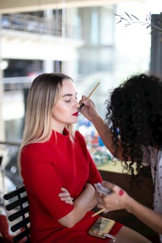 Makeup artist applying makeup to a woman seated indoors during the day, showcasing a vibrant and professional atmosphere.