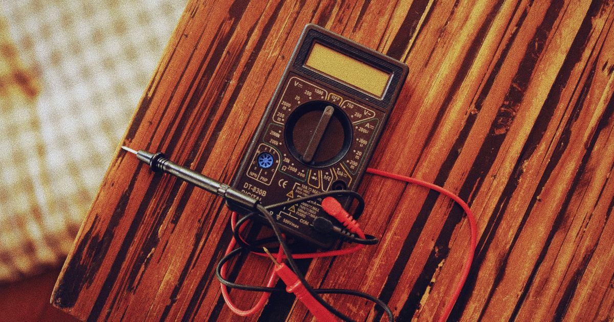Digital multimeter with red and black test leads on a weathered wooden table beside a soldering iron