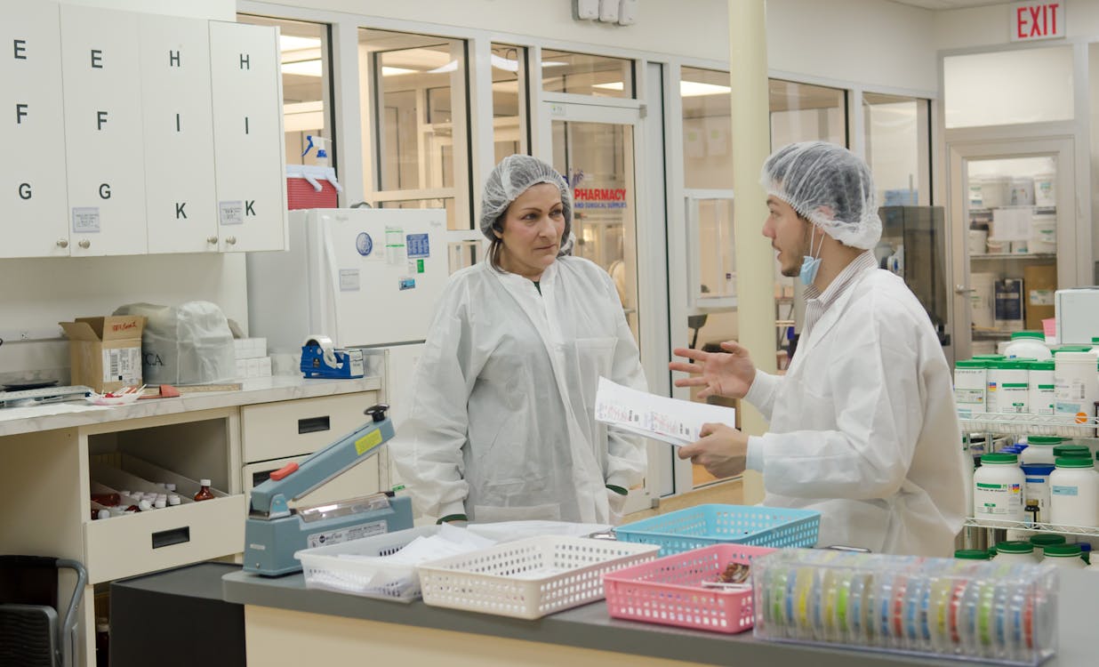 Free Concentrated laboratory workers wearing white robes and hats standing with papers in contemporary chemical room and discussing results of scientific research Stock Photo