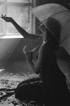 Artistic black and white photo of a woman with an umbrella in the rain by a window, creating a conceptual mood.