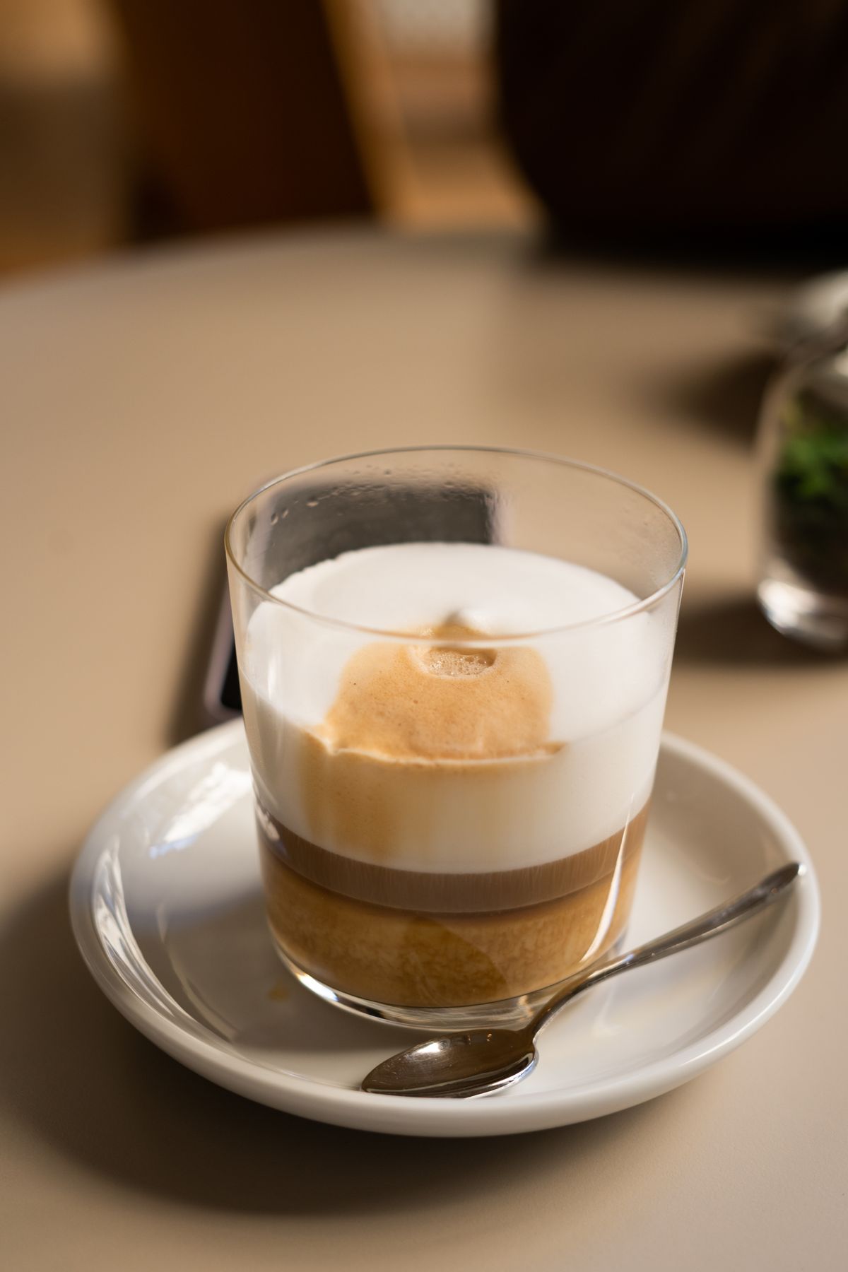 Pahar de sticlă cu strat gros de spumă de lapte peste espresso, pe farfurie albă