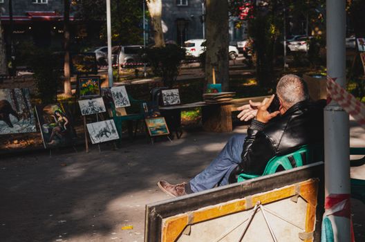 A man in a leather jacket sits on a street making a phone call, surrounded by paintings on display.