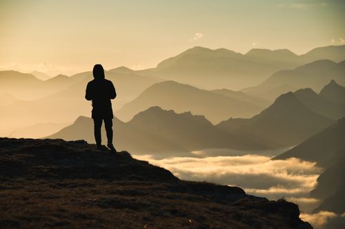 Free Silhouette of a person standing on a cliff with a stunning mountain view at dawn, surrounded by clouds. Stock Photo