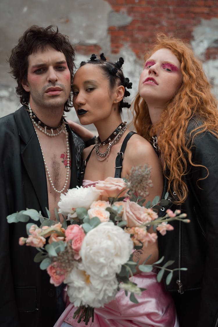 Woman In Choker Holding Bouquet Of Flowers While Standing Between Man In Black Jacket And Woman With Ginger Hair