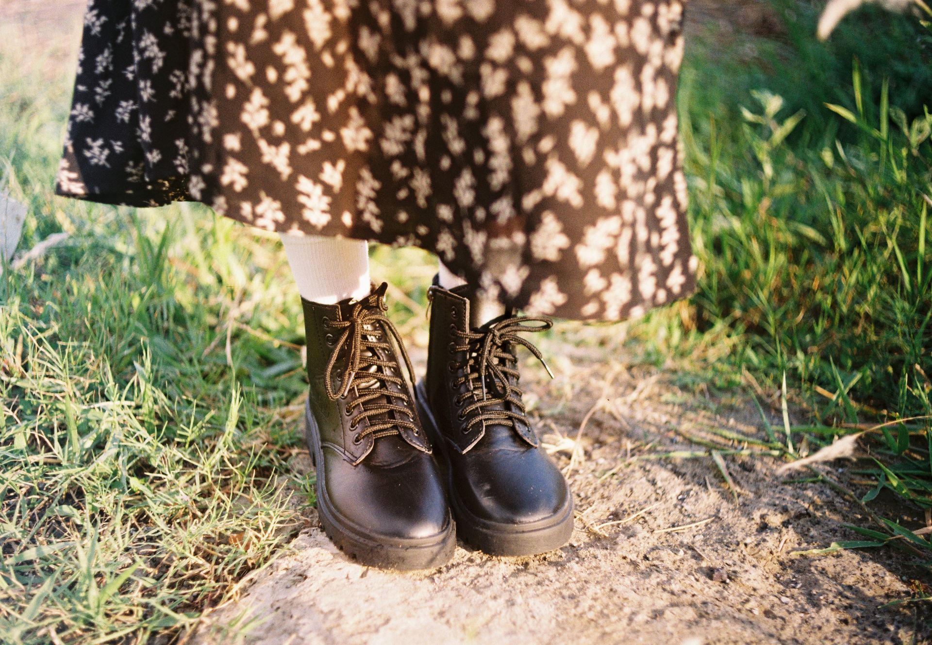 A person wearing black boots and a floral dress stands on grass in sunlight.