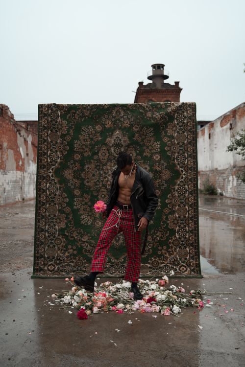 Free Bold fashion portrait of a man in plaid pants and leather jacket against a patterned rug in an urban setting. Stock Photo