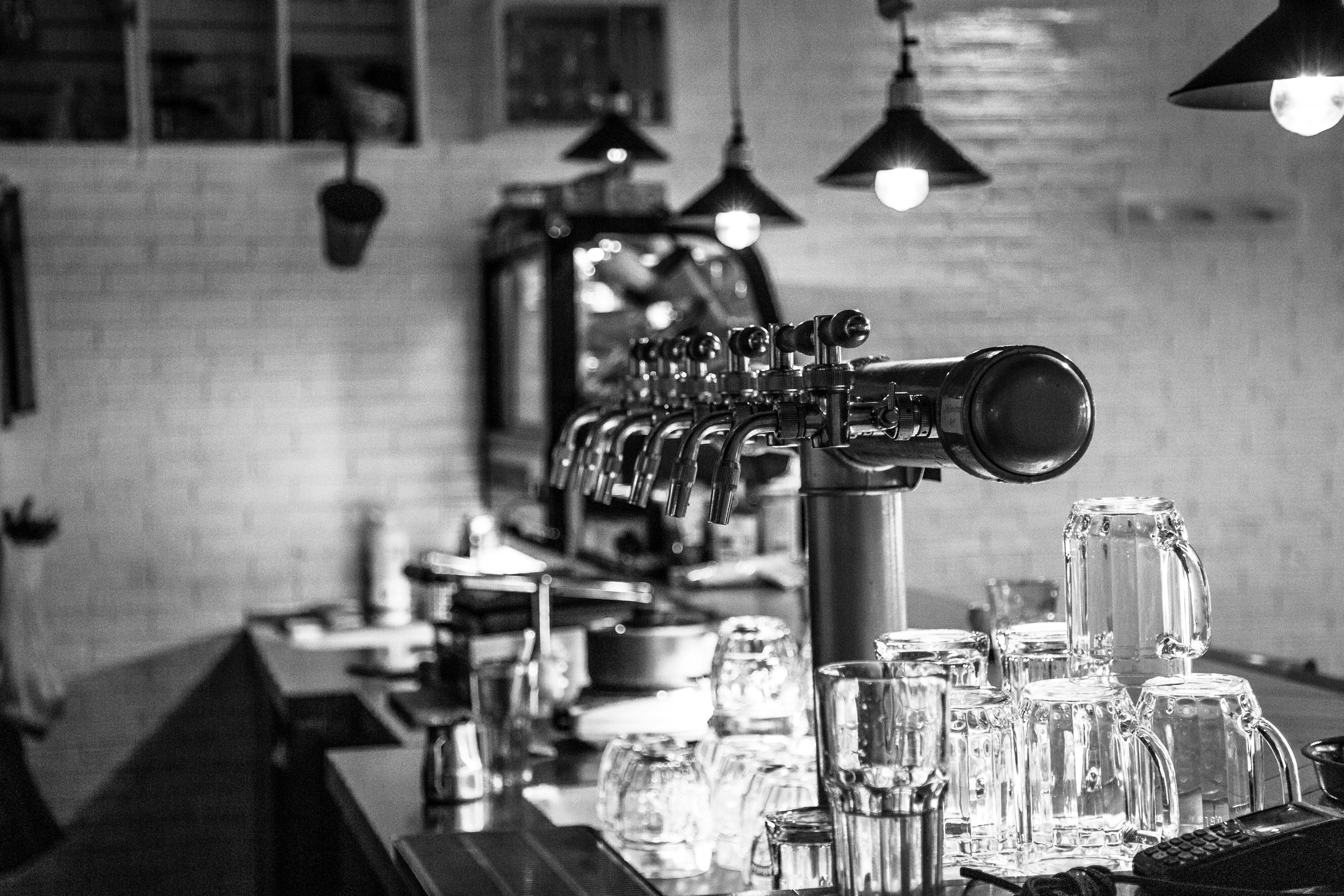 Monochrome image of a bar counter with beer taps and glassware, featuring industrial lighting.