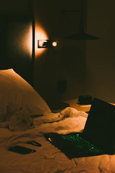A warm and cozy bedroom setting featuring a glowing laptop and ambient lighting, ideal for relaxation.