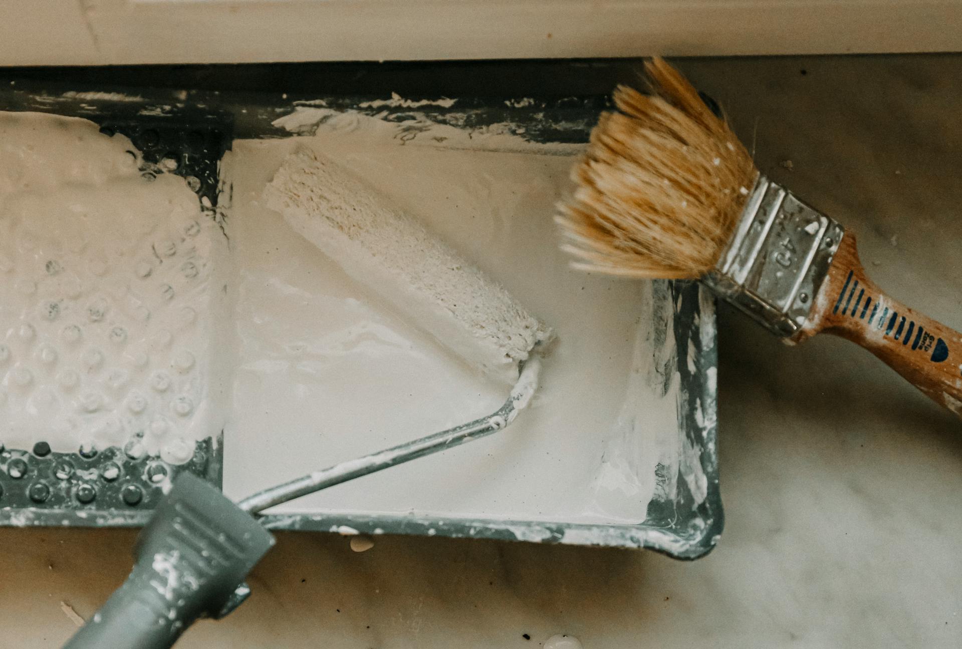 Overhead view of a paint roller and brush in a tray with white paint. Perfect for DIY and home renovation themes.