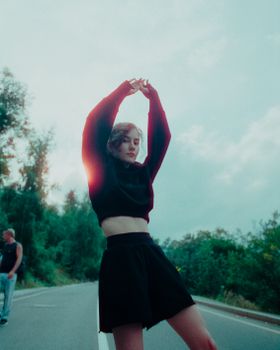 Woman in a black outfit poses on an open road, arms stretched upward, surrounded by greenery.