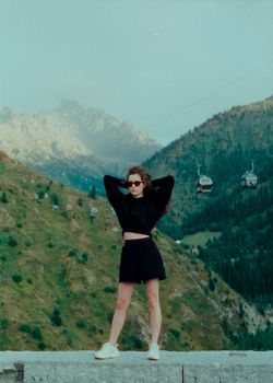 Stylish woman in black clothes poses against a mountain backdrop in Almaty, Kazakhstan.