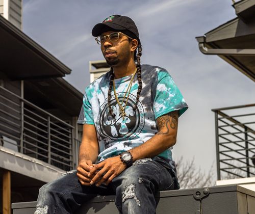 Free Man in tie-dye shirt and cap sitting outdoors in a casual urban setting. Stock Photo