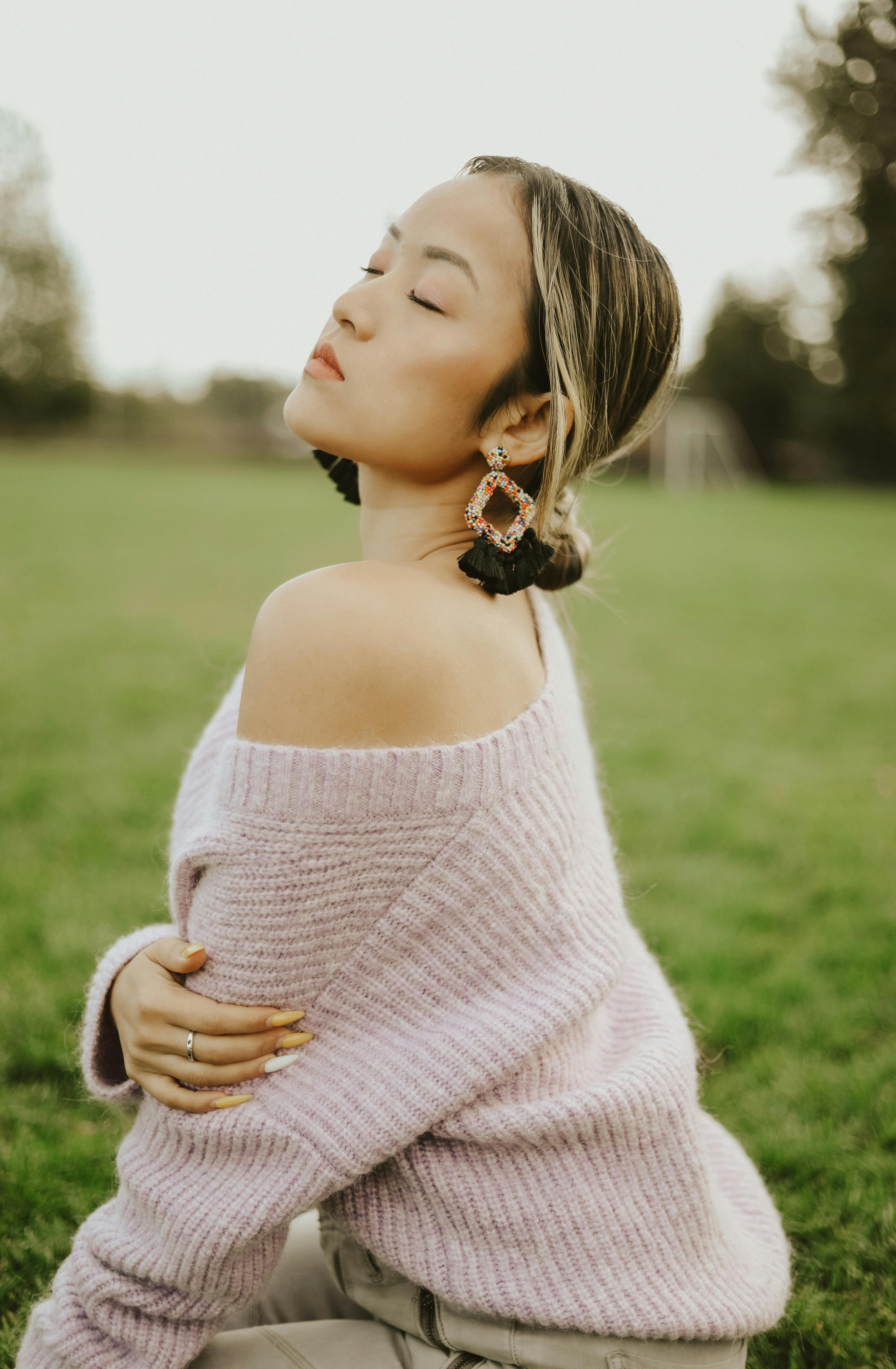 Woman sitting in a field, eyes closed, wearing a sweater and large earrings, capturing a serene moment.