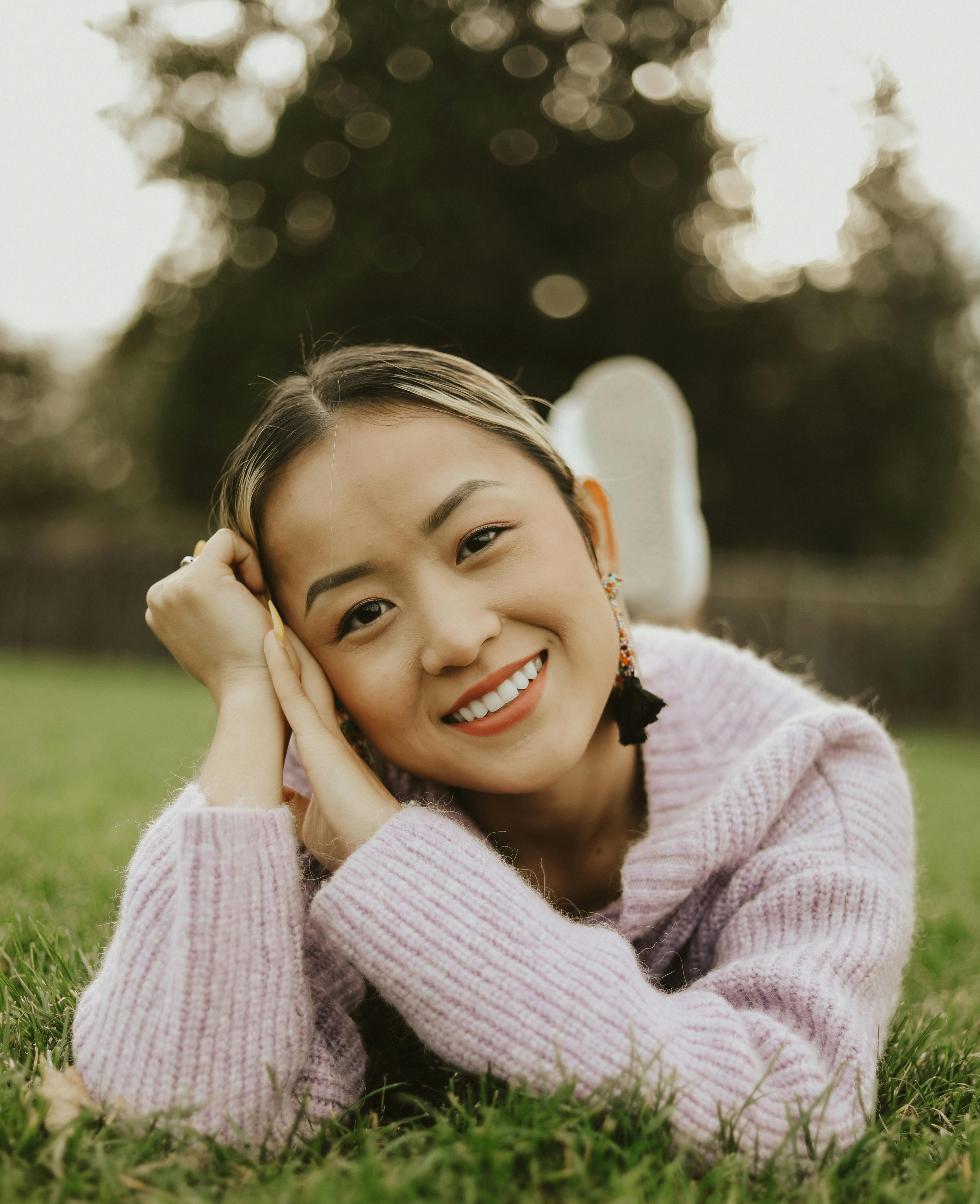 Young woman in a cozy sweater smiling while lying on grass in a serene outdoor setting.