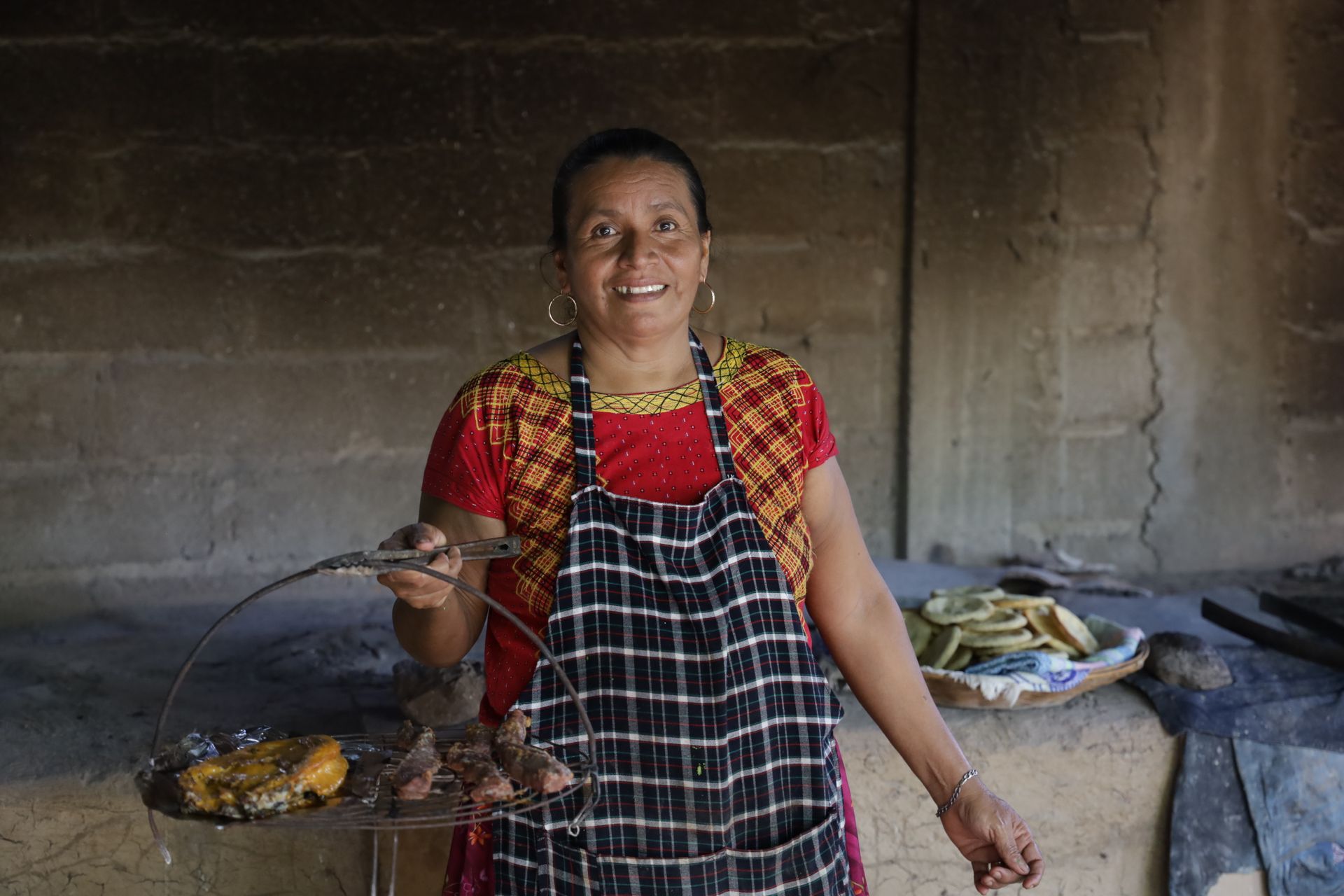 A woman in a colorful outfit grills food indoors with a smile, showcasing traditional cooking.