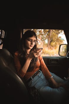 A young woman wearing glasses and casual clothing relaxes in a car, using her phone on a sunny day.