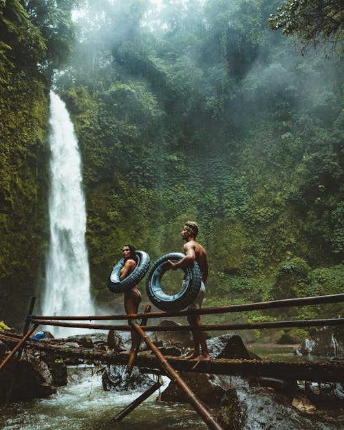 滝の近くの茶色の木の橋に黒いインフレータブルプールフロートを運ぶ二人