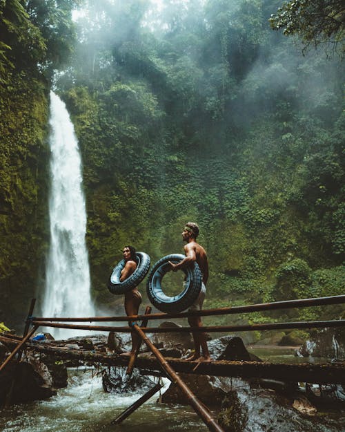 Two Person Carrying Black Inflatable Pool Float on Brown Wooden Bridge Near Waterfalls