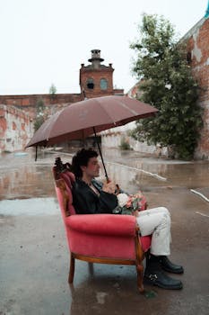 A man sits on a pink armchair holding an umbrella in a rainy urban environment, showcasing a unique juxtaposition of elements.
