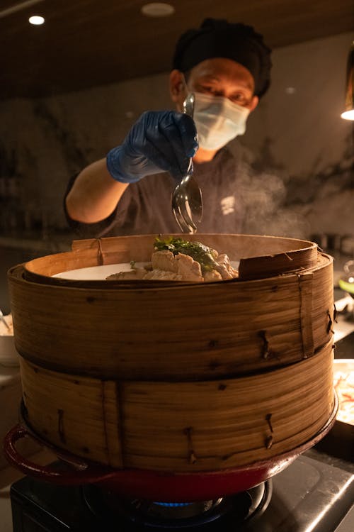 Man Wearing a Glove Holding a Spoon Over a Bamboo Steamer