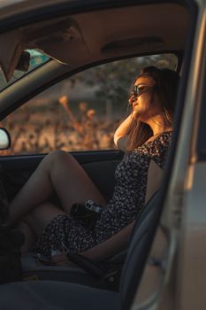 Woman sitting in car, enjoying sunset through open door, wearing floral dress and sunglasses.