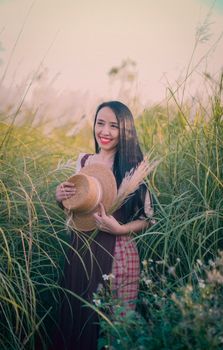 Asian woman in a dress holding a hat and pampas grass while standing in a tall, green field.