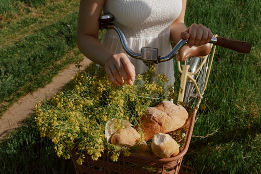 A woman in a white dress with a bicycle basket filled with flowers and bread outdoors.