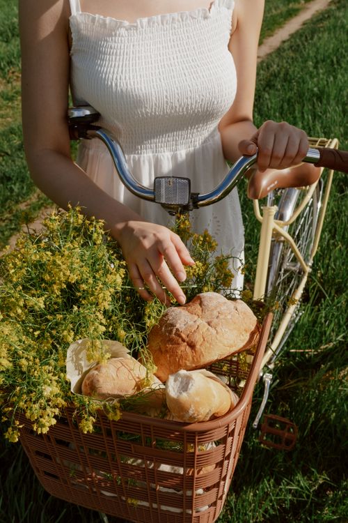 Free A close-up of a woman holding a bicycle with bread and flowers in the basket, enjoying an outdoor setting. Stock Photo