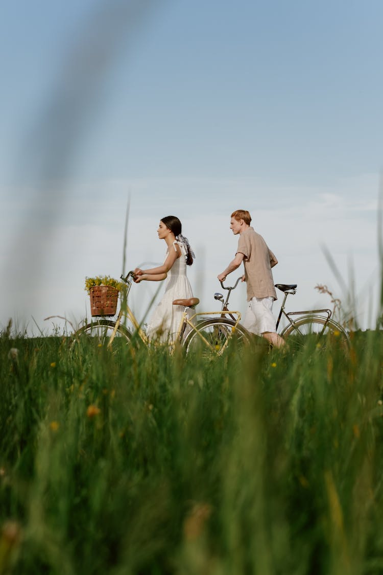 A Man And Woman Walking On The Field While Carrying Their Bicycle