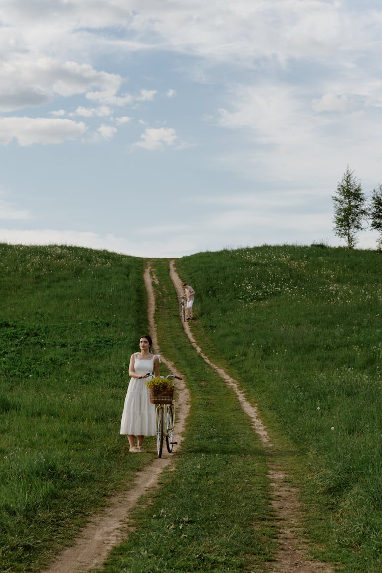 A Woman In White Dress Standing On The Field While Holding Her Bike