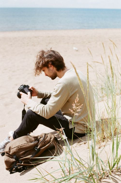 Free A young man sits on a sandy beach, examining his camera. Beach and leisure vibes. Stock Photo