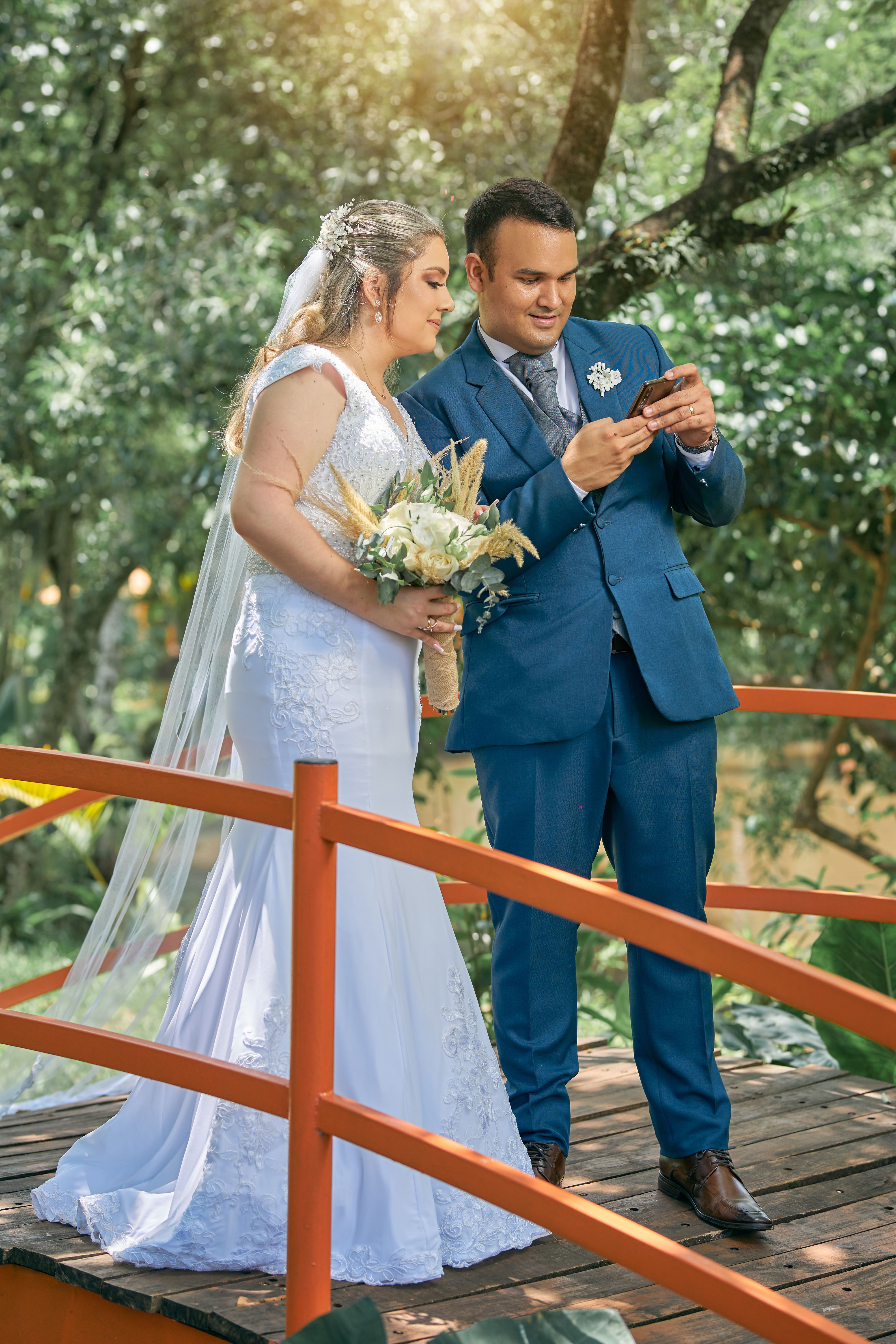 Lovely couple in wedding attire using smartphone on wooden bridge amidst lush greenery.