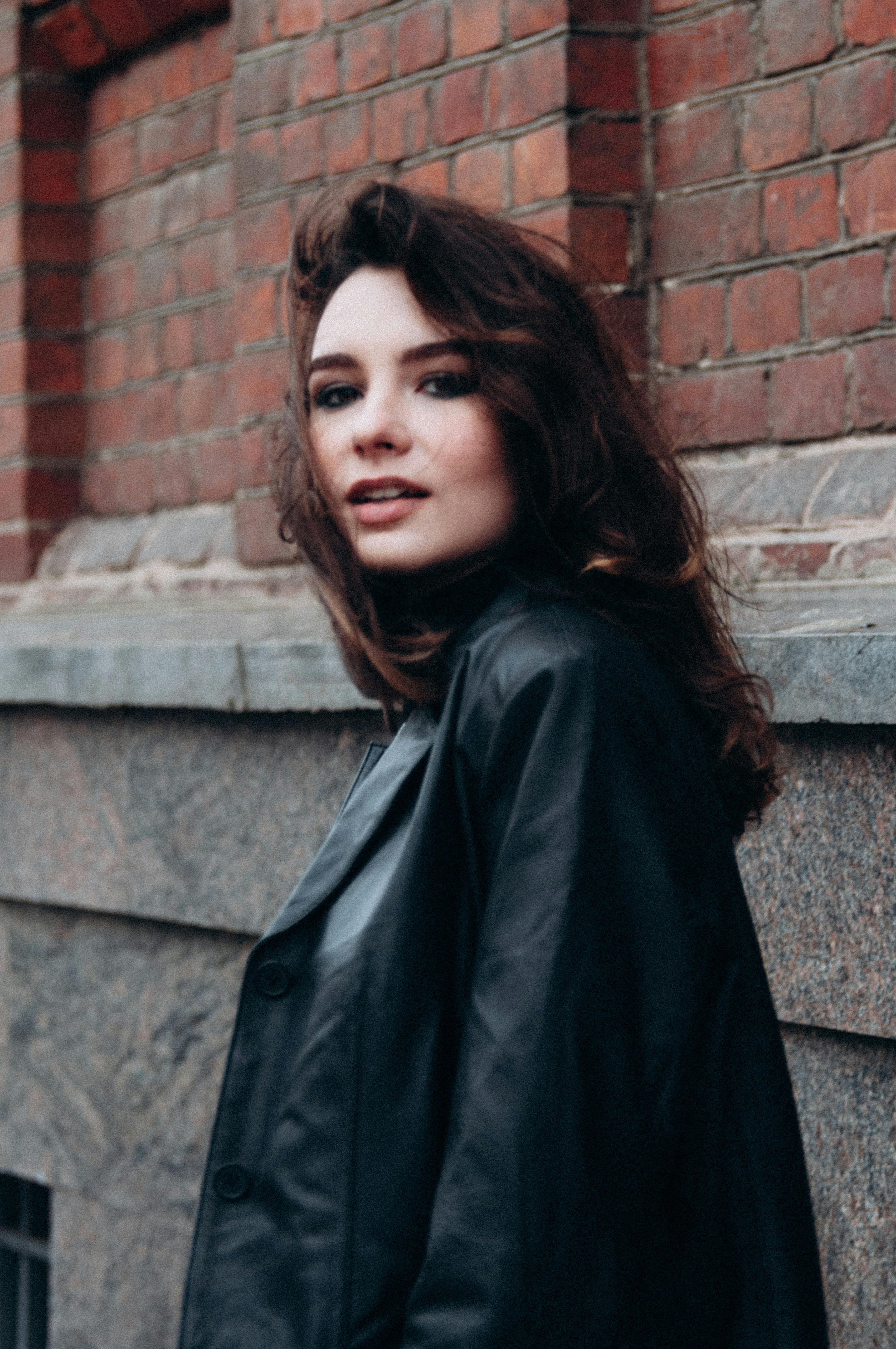 Fashionable woman in leather coat posing confidently outdoors near a brick wall.