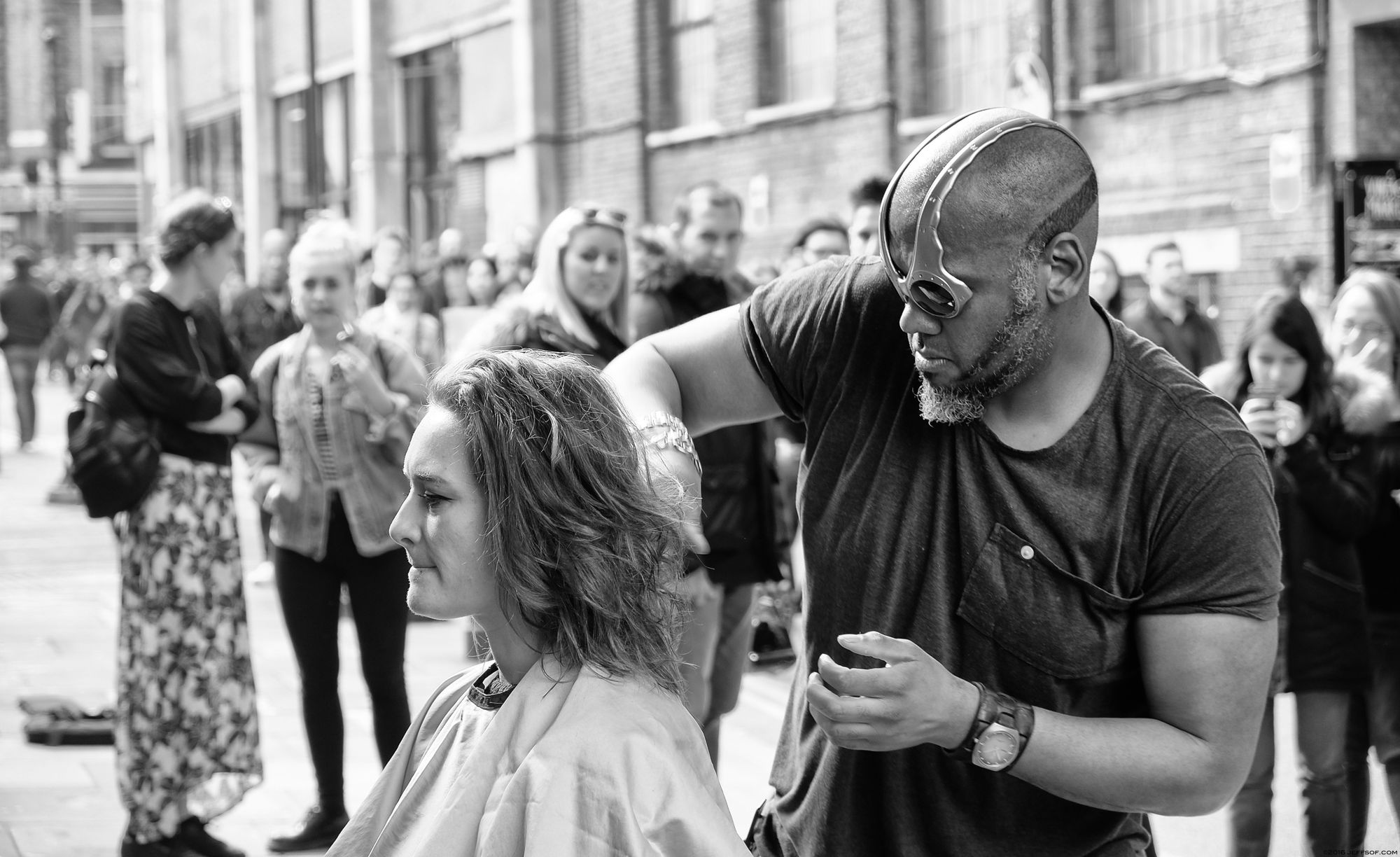 Black-and-white street photograph of a hairdresser cutting a woman's hair among a group of people in Greater London