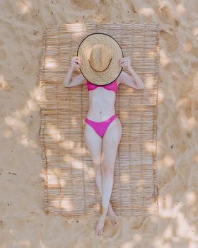 Woman in a pink bikini relaxing on a sandy beach in Caraíva, Brazil, hiding face with a straw hat.