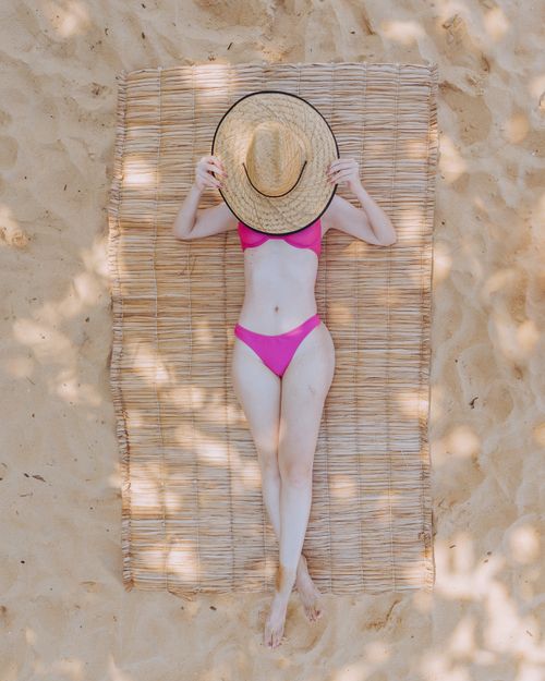 Free Woman in a pink bikini relaxing on a sandy beach in Caraíva, Brazil, hiding face with a straw hat. Stock Photo