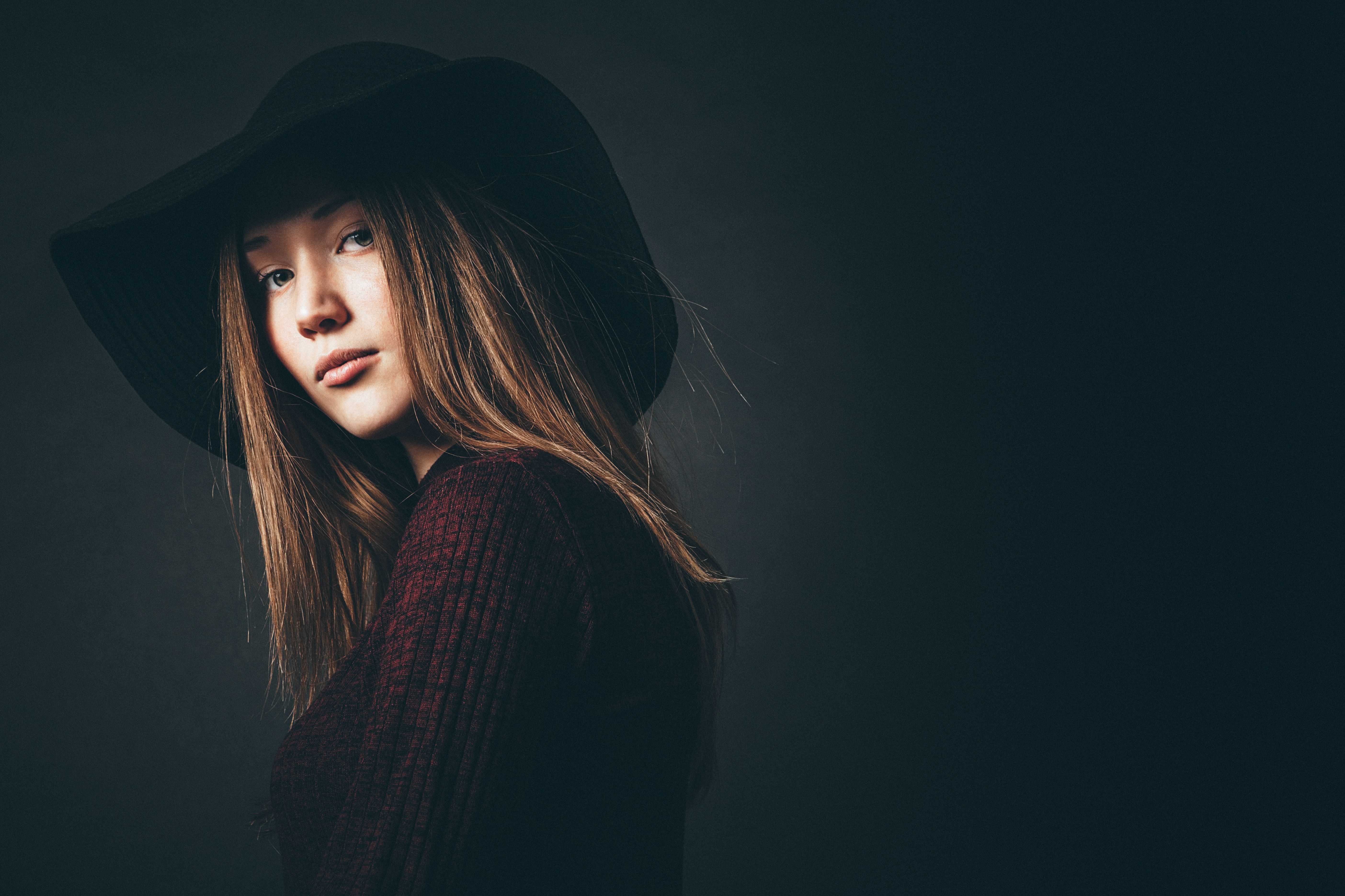 Studio portrait of a young woman wearing a black hat against a dark background, showcasing elegance and style.