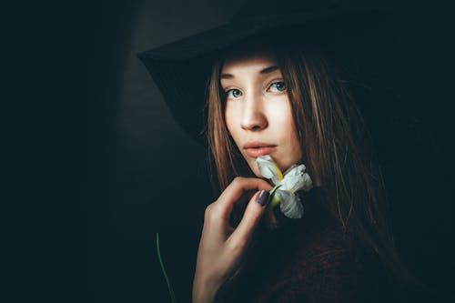 A Headshot of Young Female Holding a Flower Head, Looking at Camera and Wearing a Hat 