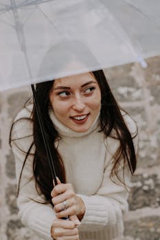 A young woman in a cozy sweater smiles while holding a transparent umbrella outdoors.