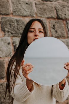 Woman holding a round mirror, reflecting the sea, against a stone wall backdrop.
