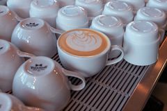 Close-up of latte art in a white cup surrounded by stacked mugs in a cozy cafe.