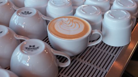 Close-up of latte art in a white cup surrounded by stacked mugs in a cozy cafe.