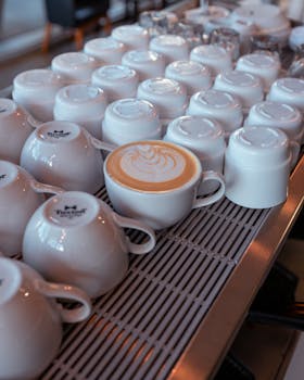 Close-up of latte art in a white cup surrounded by stacked mugs in a cozy cafe.