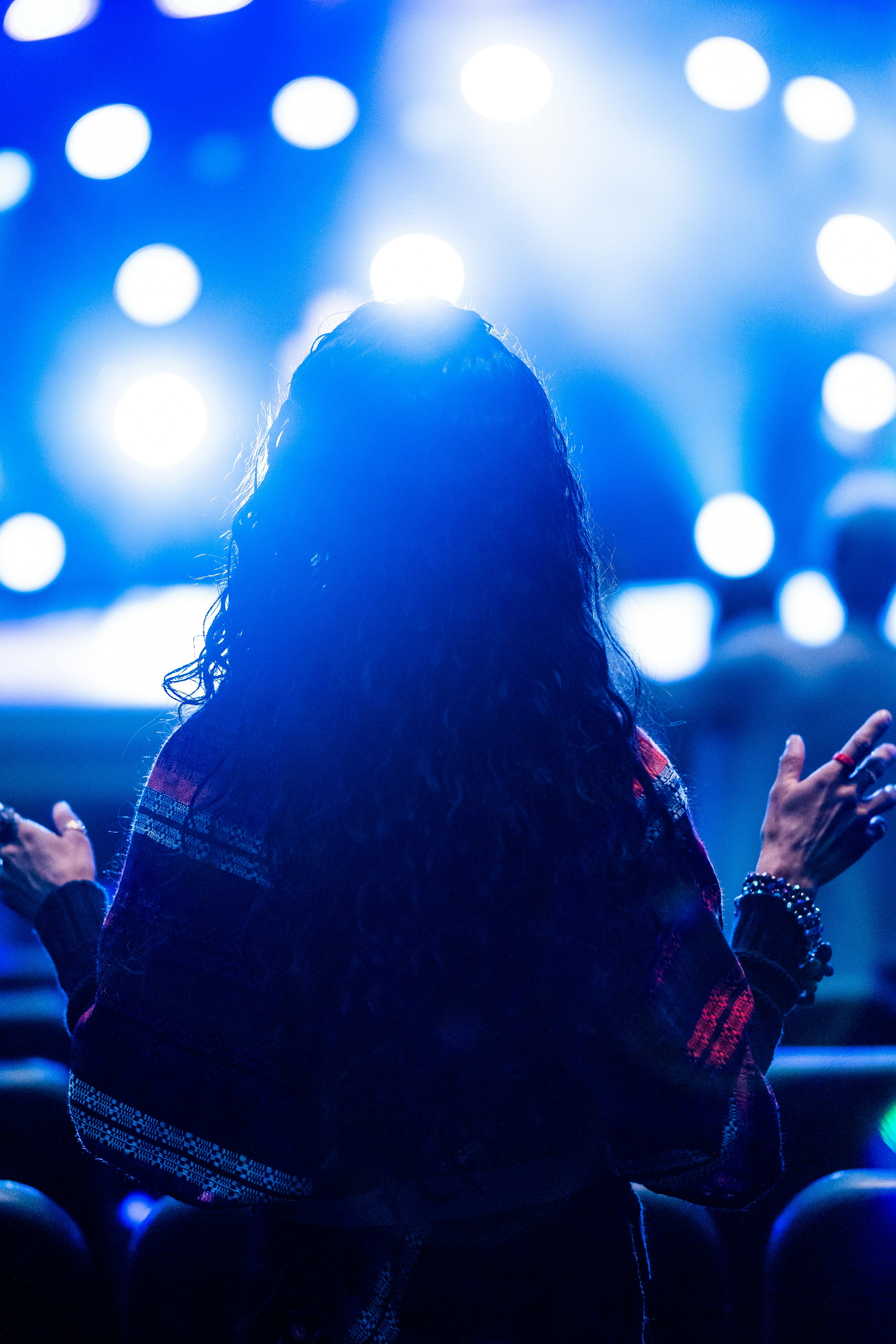 A woman raises her hands in prayer under bright, bokeh lights at a nighttime event, creating a spiritual ambiance.