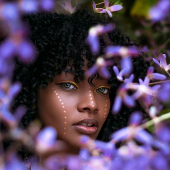 Portrait of a beautiful woman with Afro hair framed by vibrant flowers, showcasing expressive makeup and natural beauty.