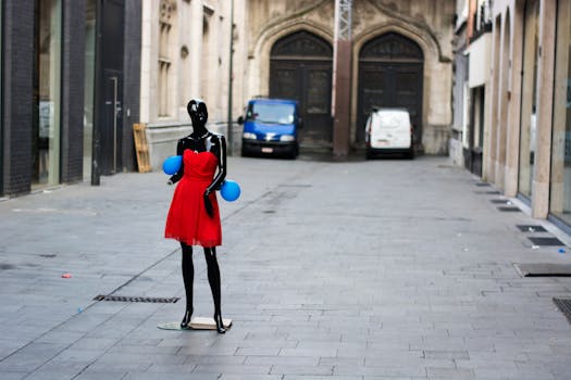 A stylish mannequin in a red dress with blue balloons on a cobblestone city street.