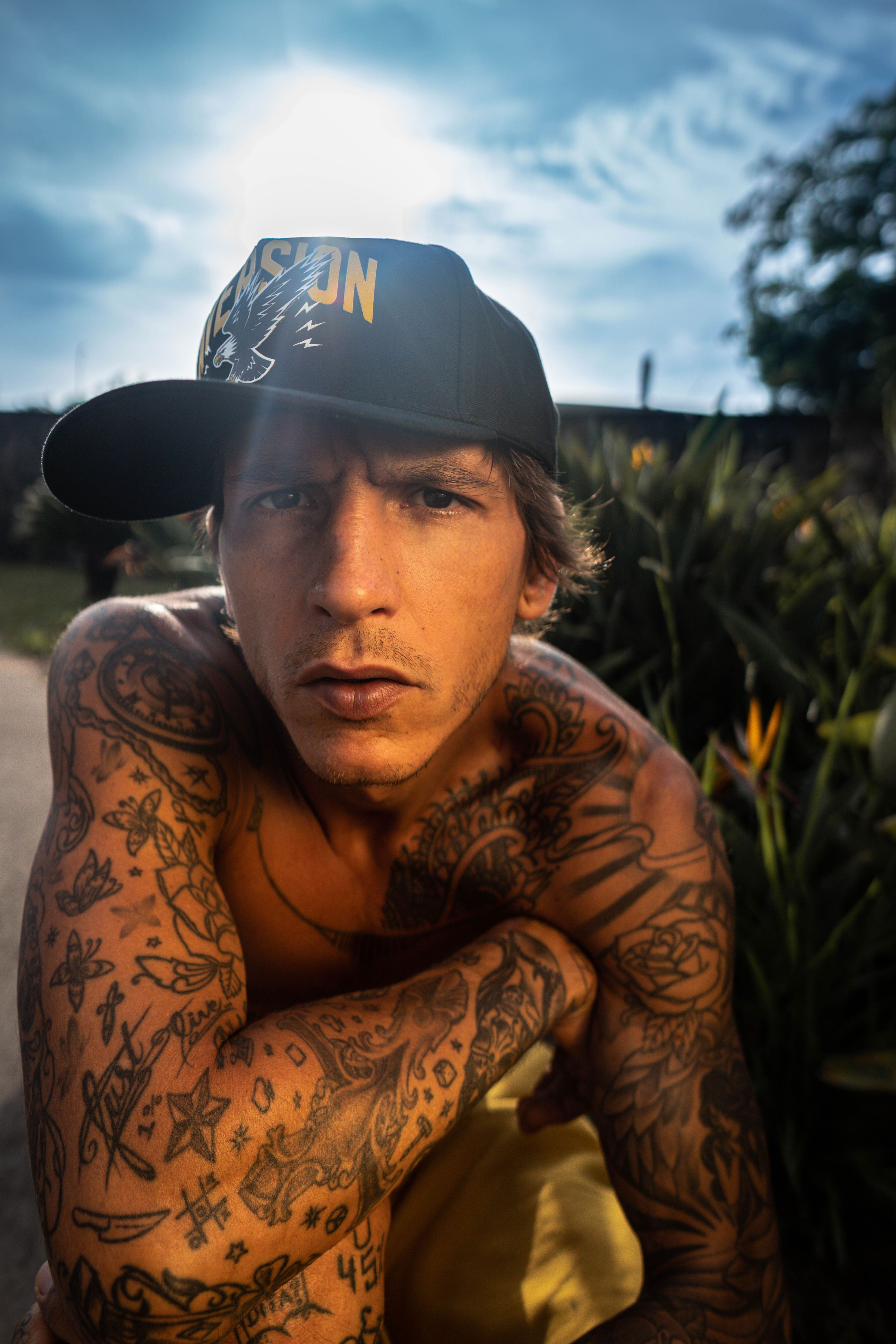 Shirtless man with tattoos and cap, looking serious outdoors in Brazil.