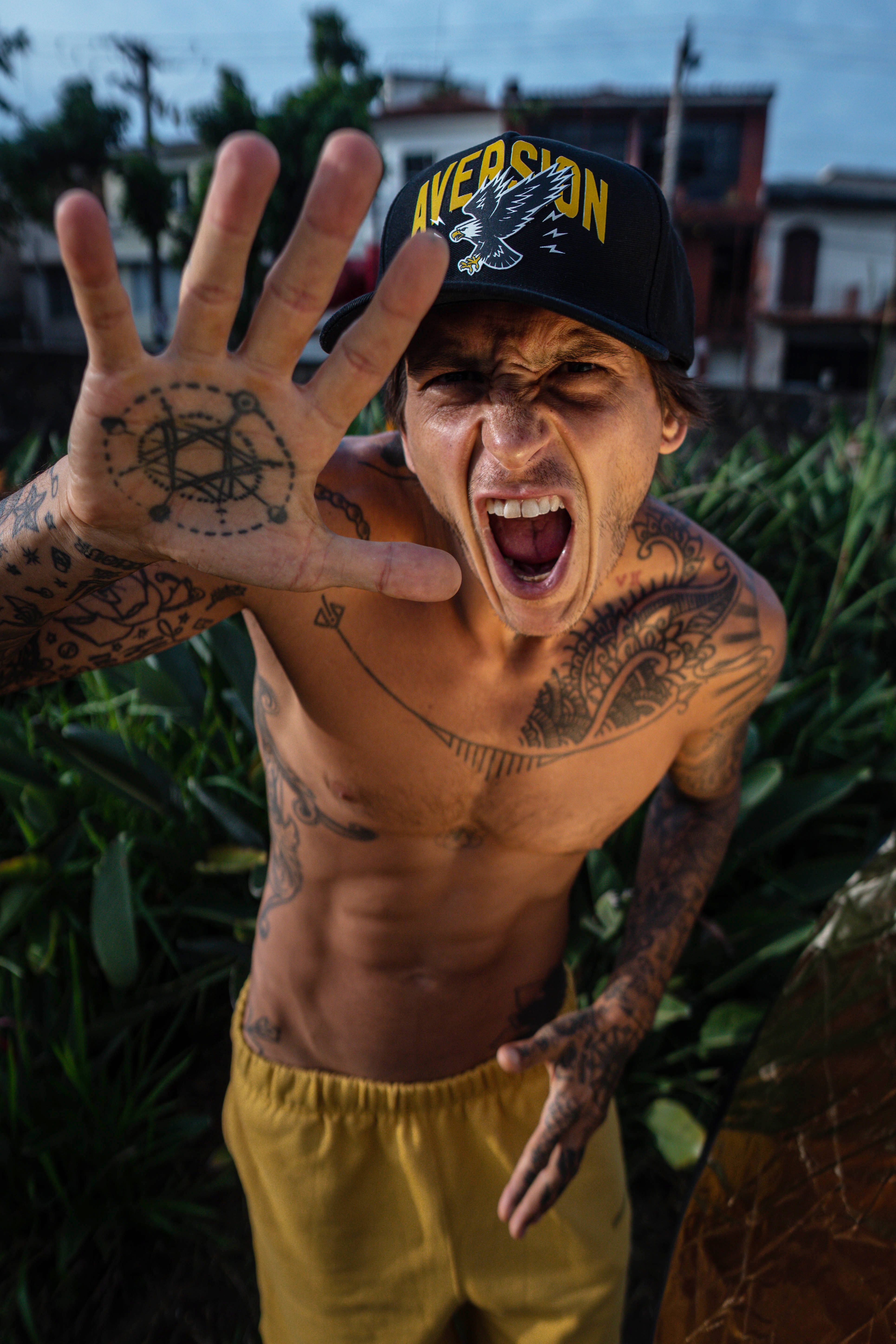 Shirtless man with tattoos and a cap yelling energetically outdoors in São Paulo.