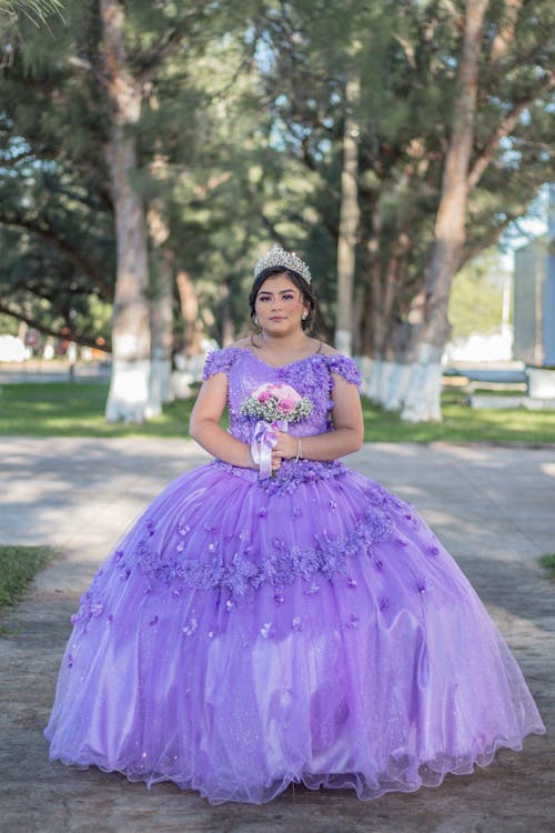 Woman in Purple Gown Holding a Bouquet of Flowers while Standing in the Park