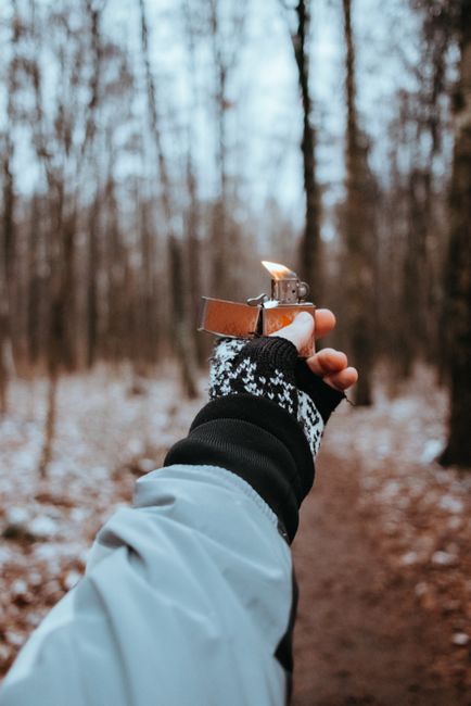 A gloved hand holds a lit lighter in a snowy forest during the winter.